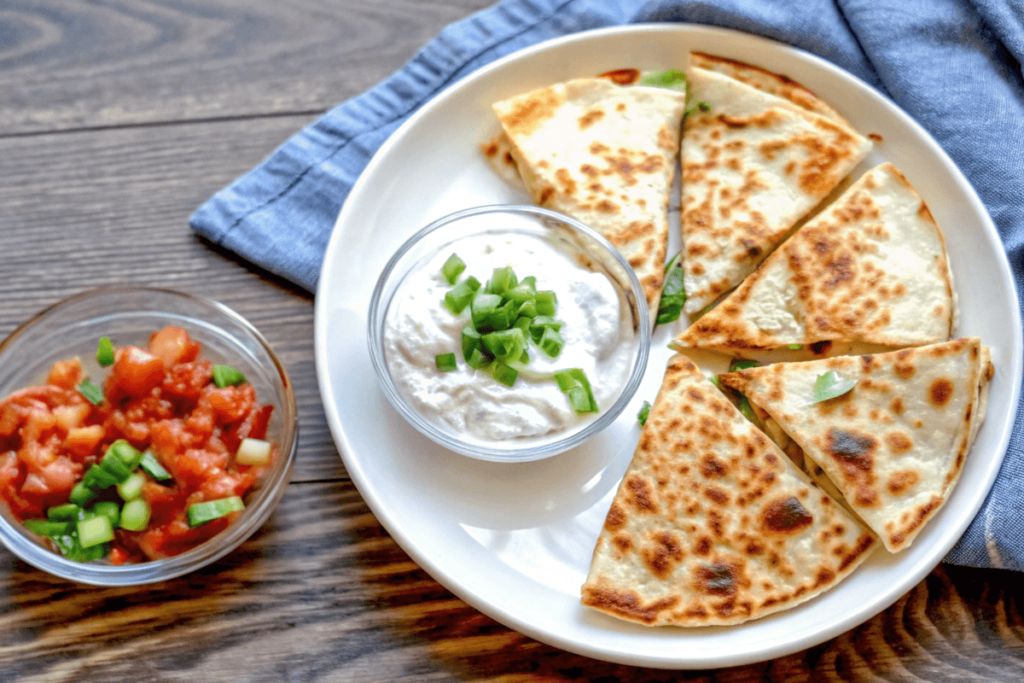 Sliced golden chicken quesadillas served with Greek yogurt, salsa, and guacamole alongside packed meal prep containers.