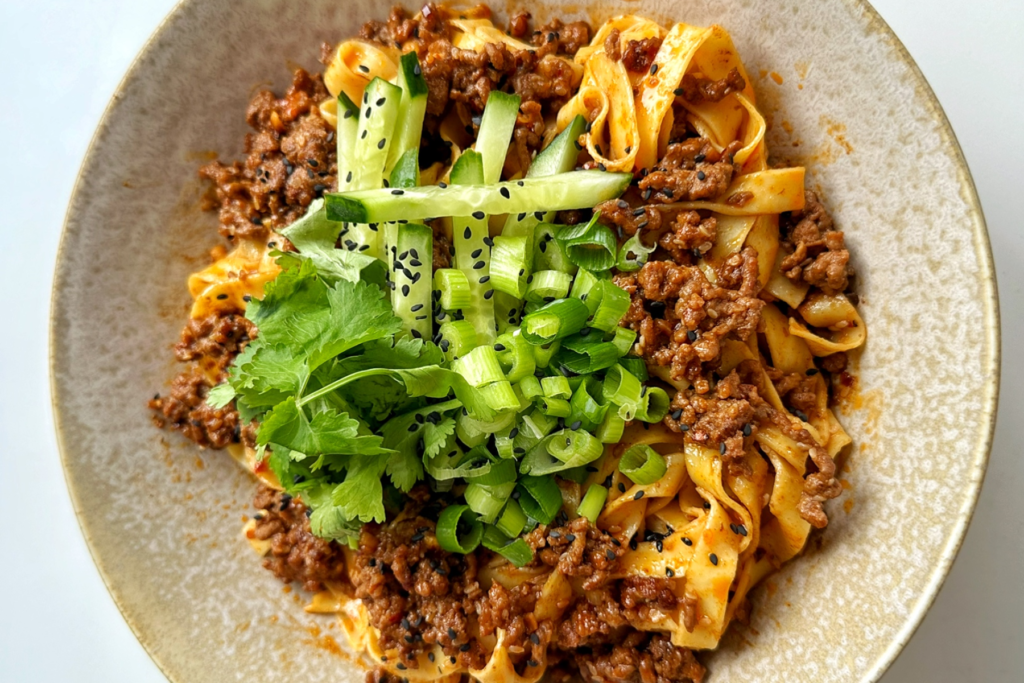 A steaming bowl of egg noodles topped with seasoned ground beef, fresh parsley, and grated cheese, served on a rustic wooden table.