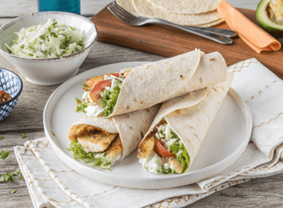 "A vibrant fish burrito filled with grilled fish, fresh vegetables, seasoned rice, and a drizzle of creamy sauce, served with guacamole and tortilla chips on a rustic wooden table.